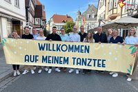 Organisatoren und Sponsoren stehen auf der Rathausstraße. Sien haben ein großes Banner in der Hand, auf dem zu lesen ist: "Willkommen auf meinem Tanztee".